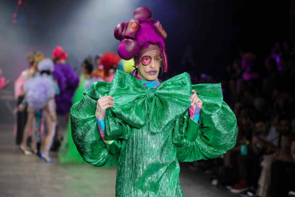 Na imagem com cor, uma pessoa branca desfilando com um vestido verde e pessoas no fundo - Metrópoles