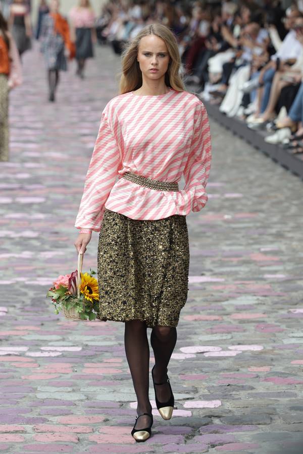 Na passarela, modelo segura cesta com flores. Ela usa blusa com listras rosas e brancas, além de saia texturizada com tons de preto e dourado - Metrópoles