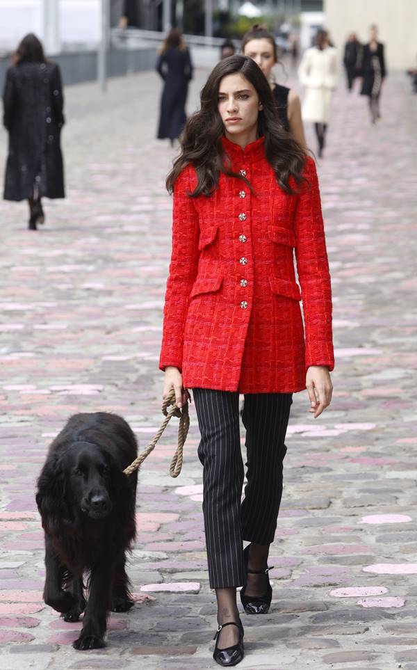Em passarela de desfile, mulher usa casaco vermelho e calça preta. Ela desfila segurando a coleira de um cachorro preto - Metrópoles