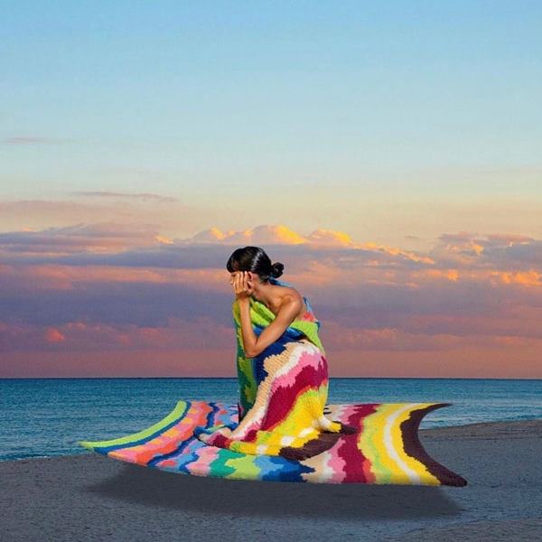 Modelo agachada em areia de praia. Ela está usando uma túnica em crochê - Metrópoles
