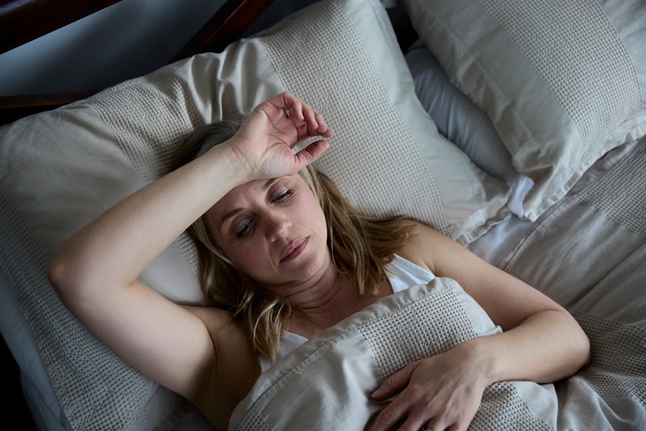 Foto van een jonge blanke vrouw die ligt met een ongemakkelijke uitdrukking, wat het gevoel van nachtelijk zweten illustreert dat wordt veroorzaakt door nierkanker - Metropoles
