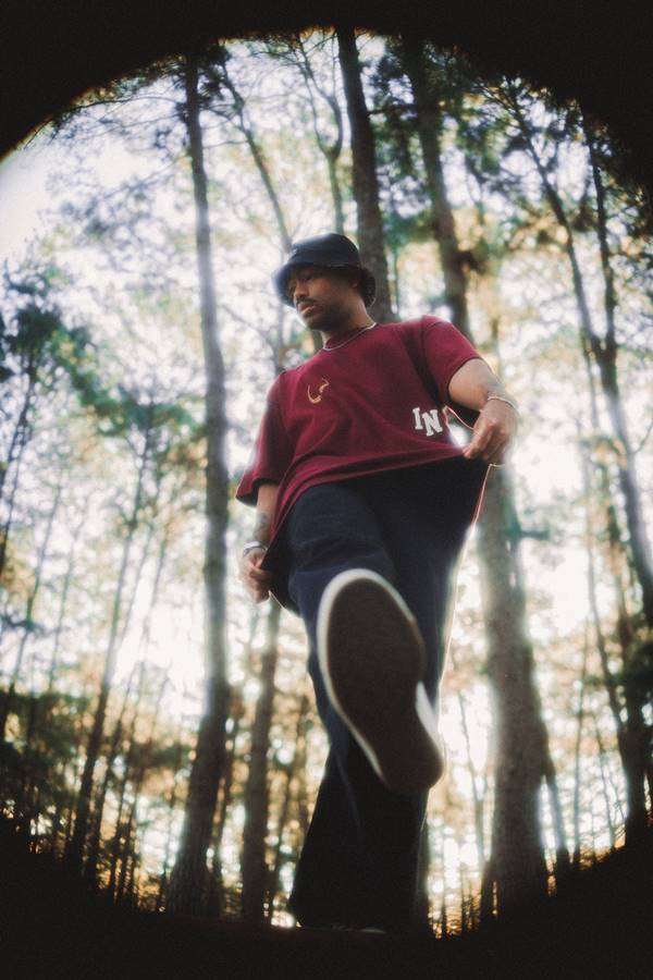 Na imagem com cor, homem negro posa para foto em parque usando camiseta vinho, calça e boné preto