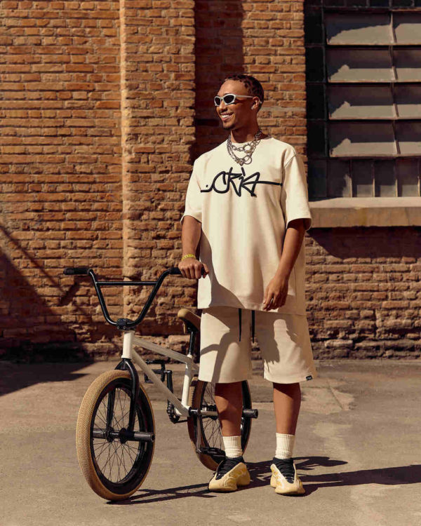 Na imagem com cor, um homem negro em pé, ao lado de uma bicicleta usando roupas claras posando para um editorial da Riachuelo - Metrópoles