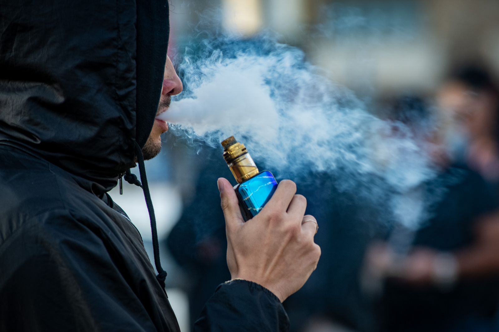 Homem branco com casaco preto e capuz fumando um cigarro eletrônico