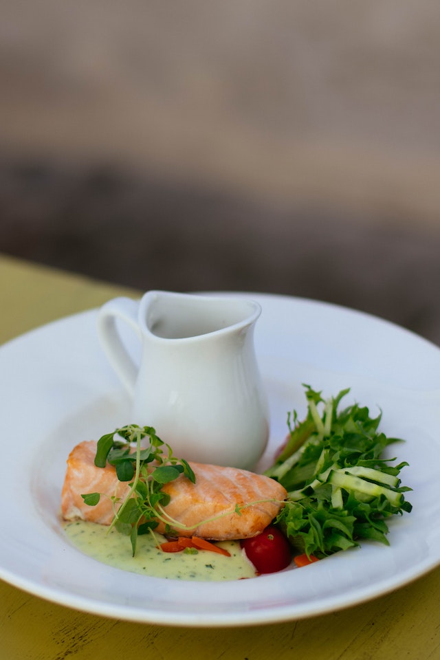 Prato de comida com uma pequena salada, uma posta de salmão e uma pequena jarra branca - Metrópoles