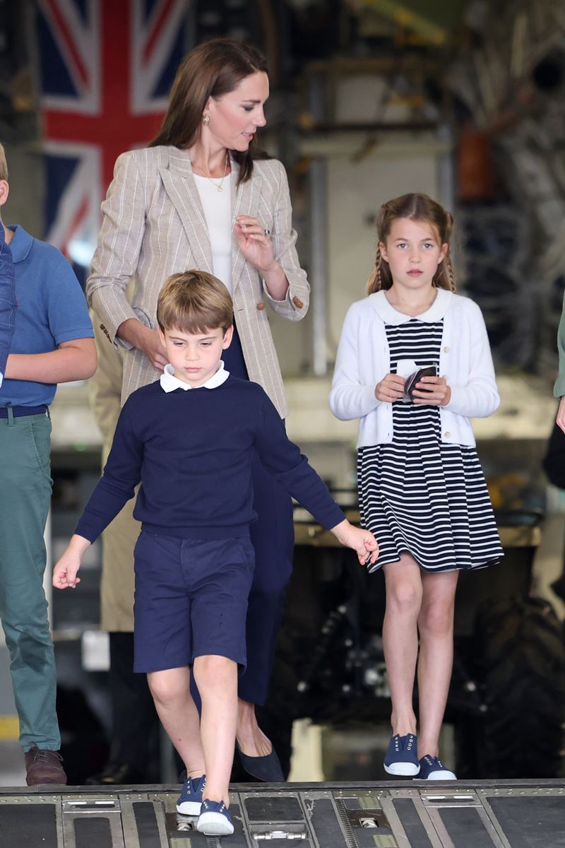 Foto colorida de menino, branco e loiro, caminhando na frente de uma mulher, branca e de cabelo castanho, e de uma menina, branca e loira. Eles saem de uma aeronave. Ao fundo, tem uma bandeira do Reino Unido - Metrópoles