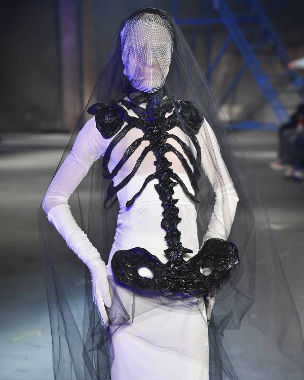 Em desfile de moda, na passarela, modelo com o rosto cobertos usa look branco com detalhes em preto que simulam um esqueleto - Metrópoles