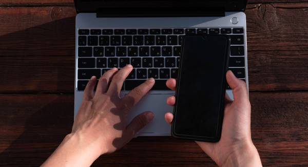 Fotografia colorida mostrando pessoa mexendo no teclado do computador com uma mão e segurando um celular com a outra-Metrópoles