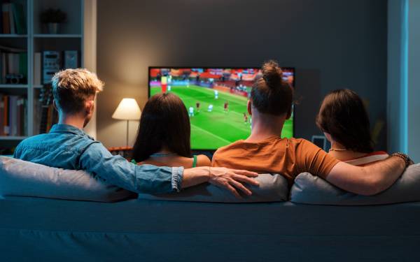 Fotografia colorida mostrando dois casais em sofá assistindo jogo de futebol na televisão-Metrópoles