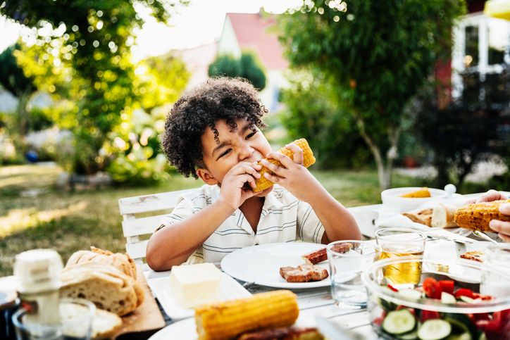 Foto de uma criança negra comendo um milho assado diante de uma bandeja com diversos alimentos