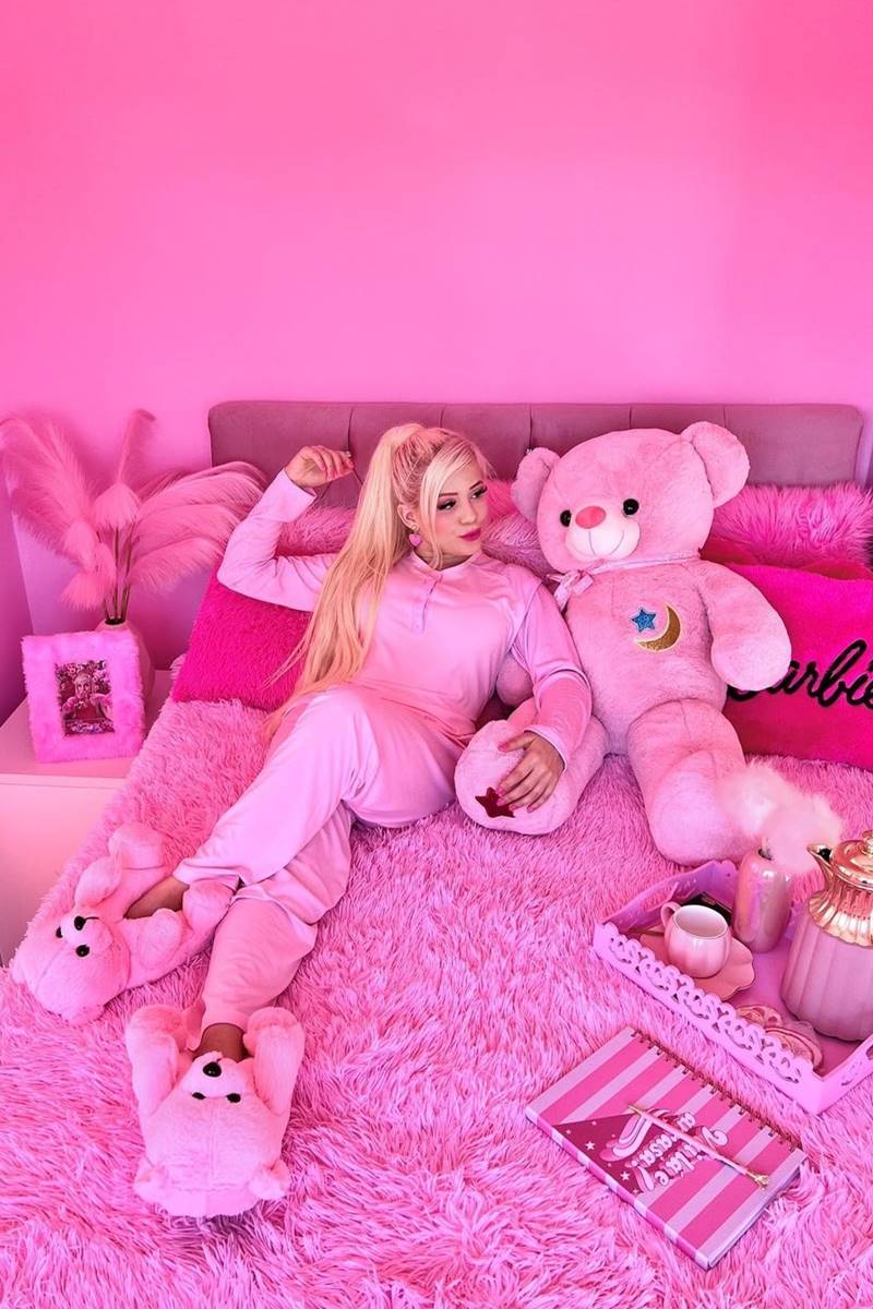 Foto colorida de mulher com roupa cor-de-rosa, deitada em uma cama, ao lado de um urso grande, também pink. Os itens decorativos do espaço são cor-de-rosa - Metrópoles