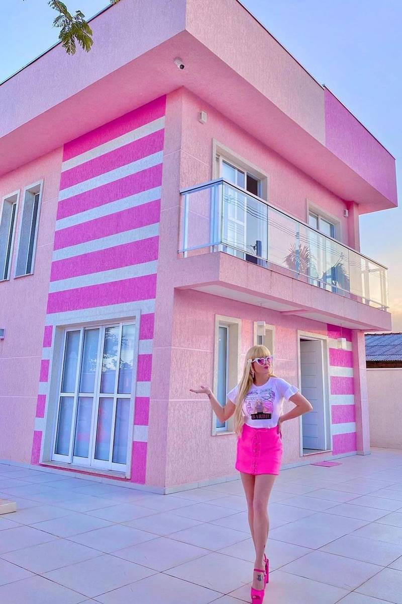 Foto de mulher loira mostrando a fachada de uma casa com paredes pintadas em cor-de-rosa - Metrópoles