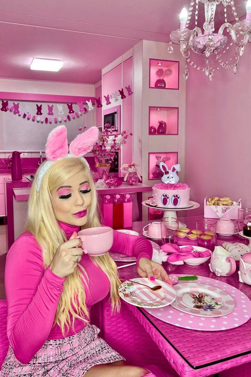 Foto colorida de mulher loira, bebendo em uma xícara cor-de-rosa, com parede pink, e decoração de cabeça de coelhinho no tom cor-de-rosa - Metrópoles