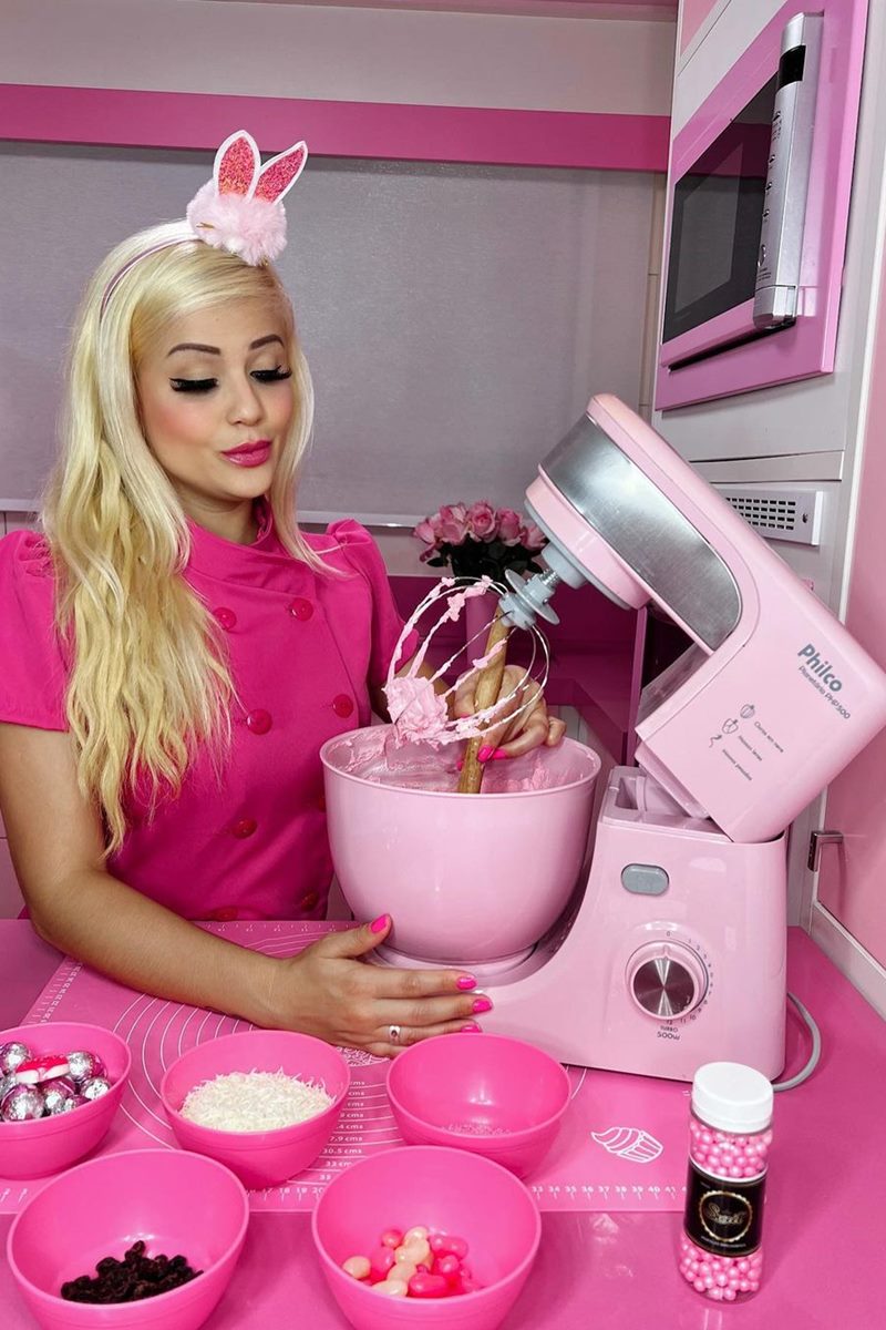 Foto colorida de mulher loira, vestida de rosa, batendo uma massa do bolo em uma batedeira cor-de-rosa, com itens decorativos na mesma tonalidade - Metrópoles