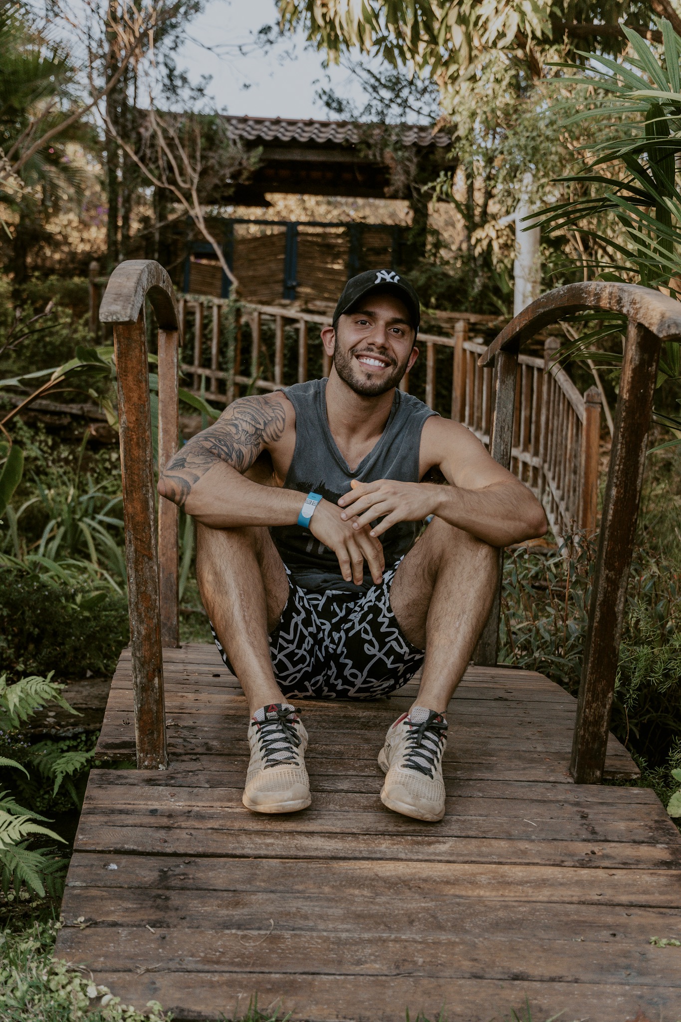 Foto colorida. Homem branco está usando boné, sentado em uma pequena ponte sorrindo. - Metrópoles 