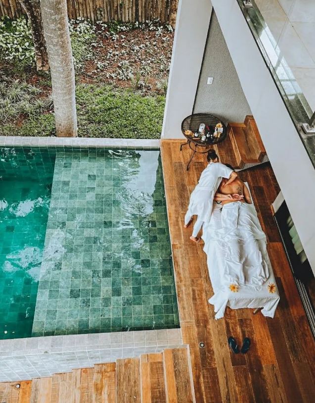 Foto colorida. Foto tirada de cima. Nela uma mulher está fazendo massagem em uma pessoa. Do lado dela está uma piscina