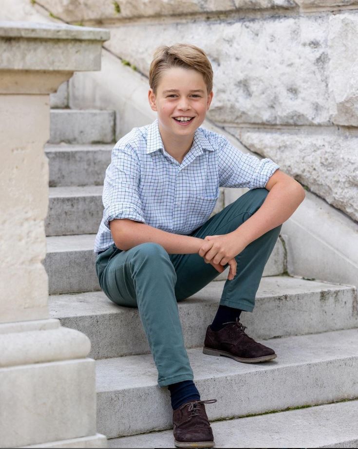 Foto Colorida. O príncipe George é um menino de 10 anos, branco e com cabelos loiros. vestindo uma camisa xadrez e uma calça azul. Com um grande sorriso no rosto, ele está sentado nas estadas do palácio e faz uma pose descontraída - Metrópoles