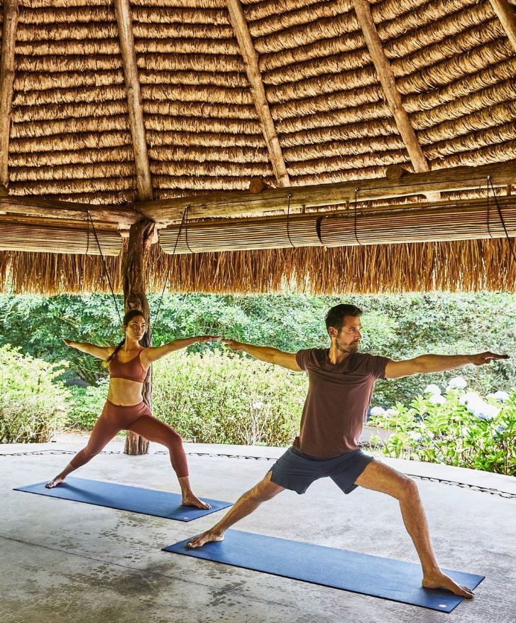 Foto colorida. Um homem e uma mulher, ambos brancos, estão fazendo movimentos de yoga - Metrópoles