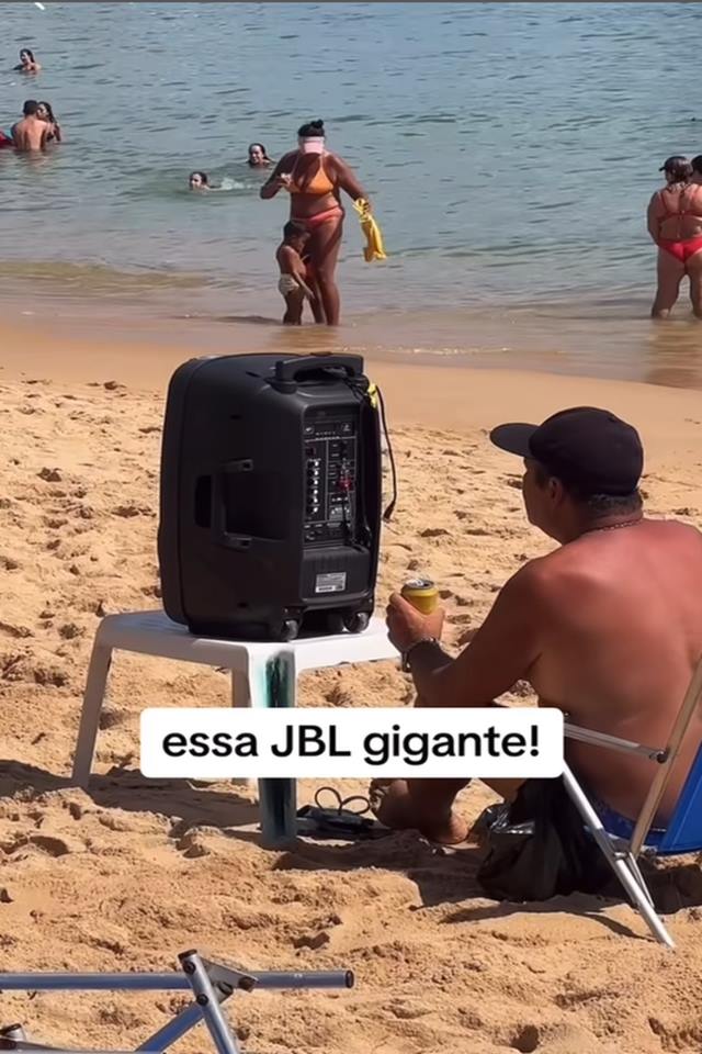 Na foto, uma caixa de som preta em cima de uma mesa na praia. Um homem branco sentado ao lado do objeto - Metrópoles 