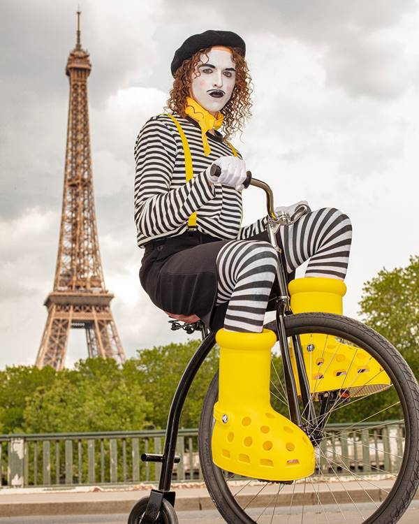 Com roupa de listrada e rosto pintado, Tommy Cash pedala com bota amarela gigante nos pés e Torre Eiffel ao fundo - Metrópoles