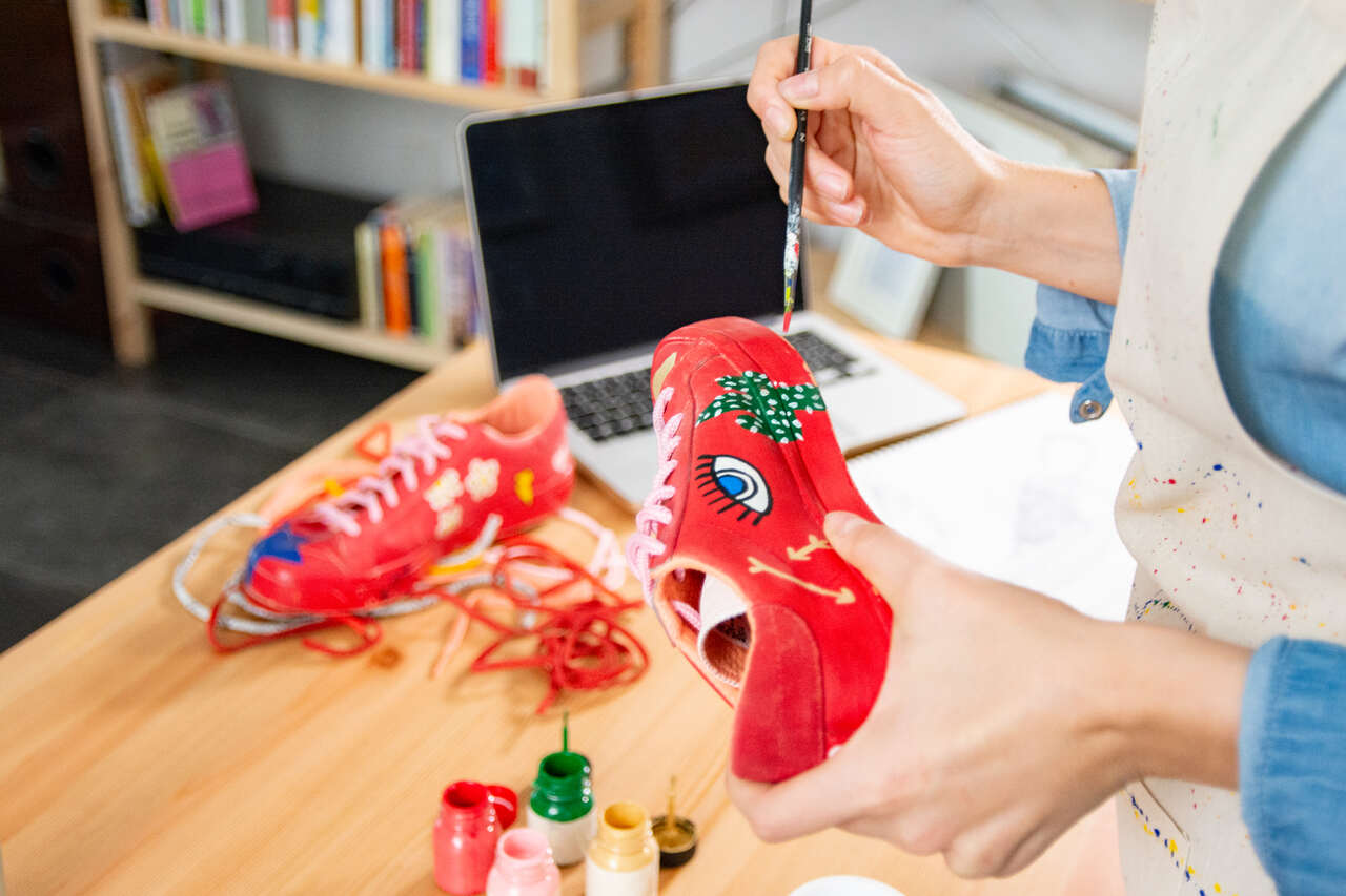 Na imagem com cor, as mãos de uma pessoa segurando um tênis vermelho e pintando-o - Metrópoles