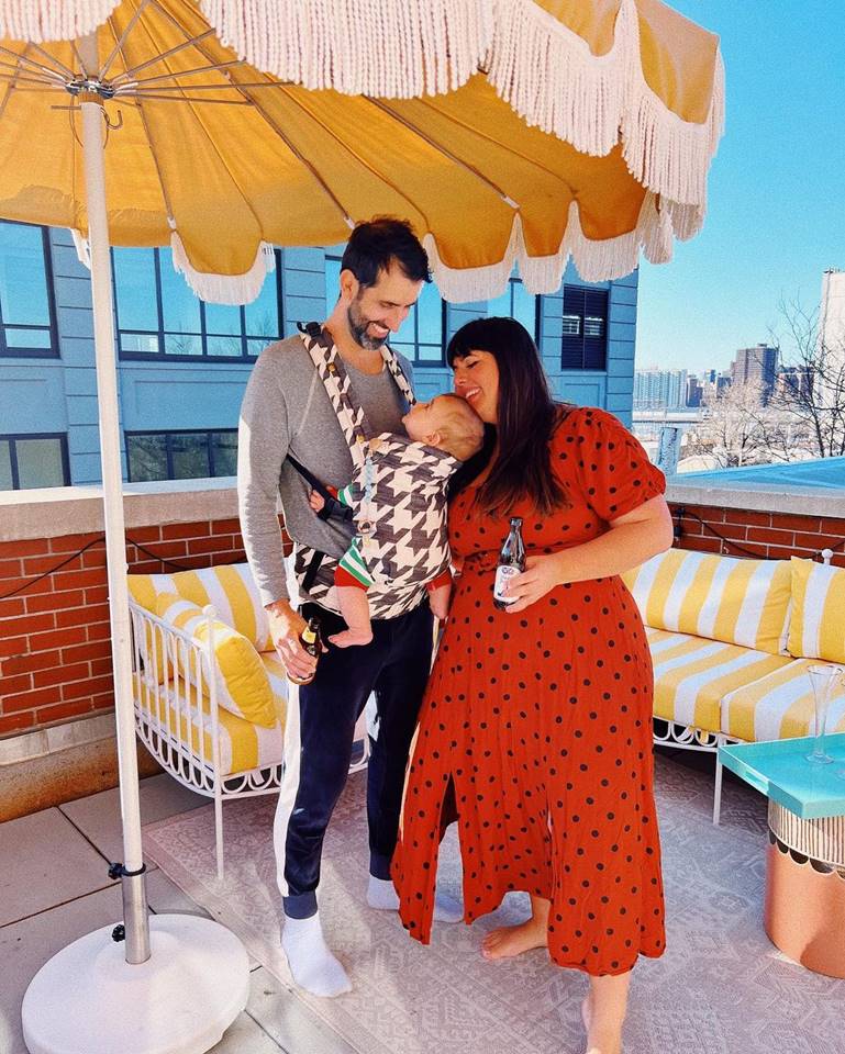 Foto colorida de casal sorrindo com um filho no colo em uma varanda com um guarda sol amarelo - Metrópoles