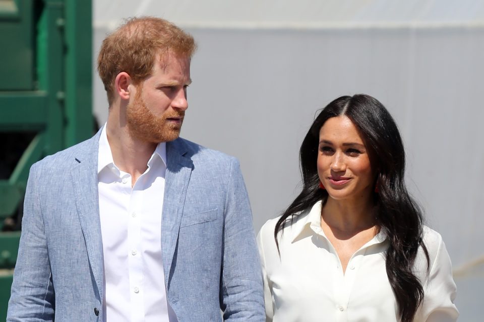 Foto colorida. Imagem mostra o casal Harry e Meghan. Ele é uma homem branco, ruivo e está usando um terno azul claro. Ela é uma mulher negra, de cabelos lisos e grandes - Metrópoles