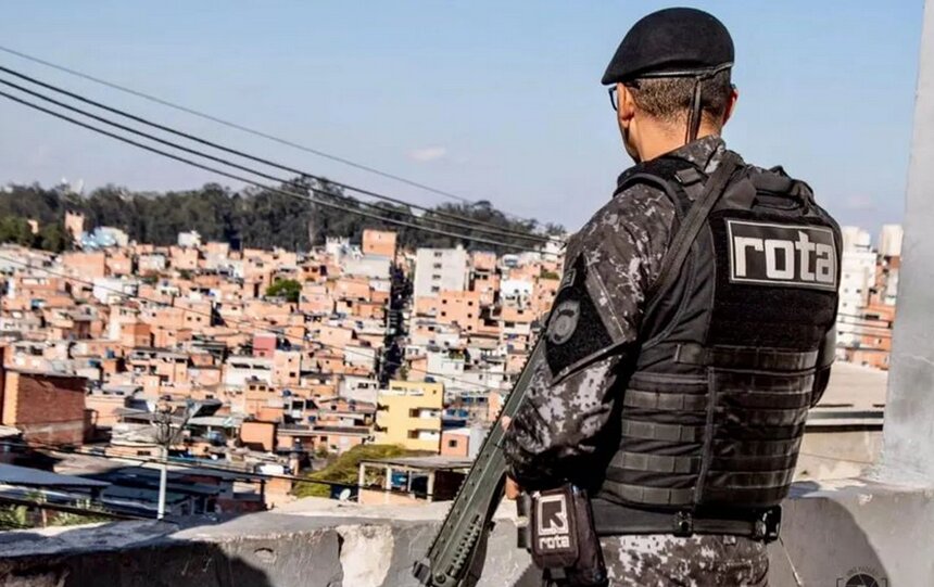 Em foto colorida policial da Rota, de costas, observa favela enquando segura um fuzil - Metrópoles