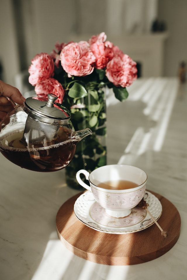 Mão despejando chá de bule transparente em xícara branca em uma mesa. Vaso com flores ao fundo - Metrópoles