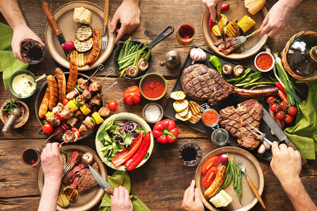 Foto colorida de pratos e tigelas com carnes, legumes, molhos, disponíveis em uma mesa de madeira. Mãos pegam os alimentos - Metrópoles