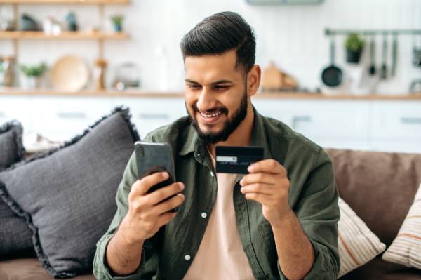 Fotografia colorida mostrando homem de barba sentado no sofá fazendo compras pelo celular com cartão de crédito-Metrópoles