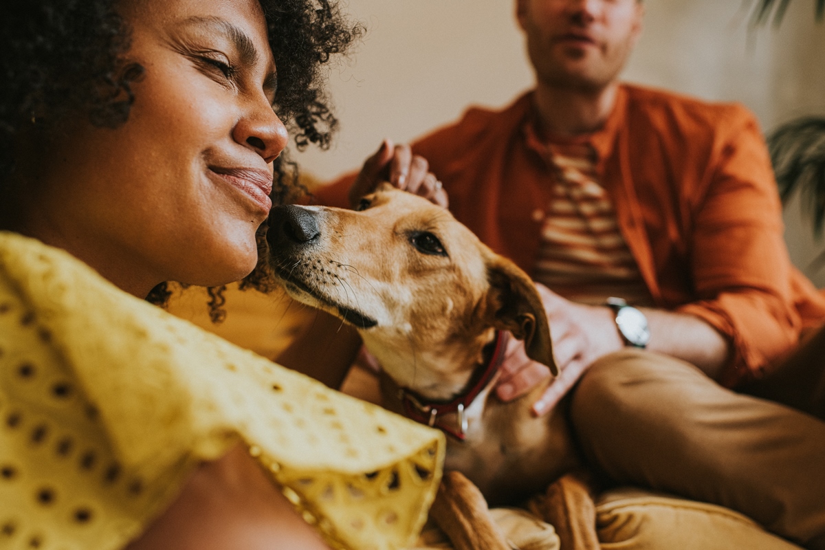 Foto colorida. Imagem mostra mulher negra sentada do lado esquerdo recebendo carinho do cachorrinho. No fundo, tem um homem sentado - Metrópoles