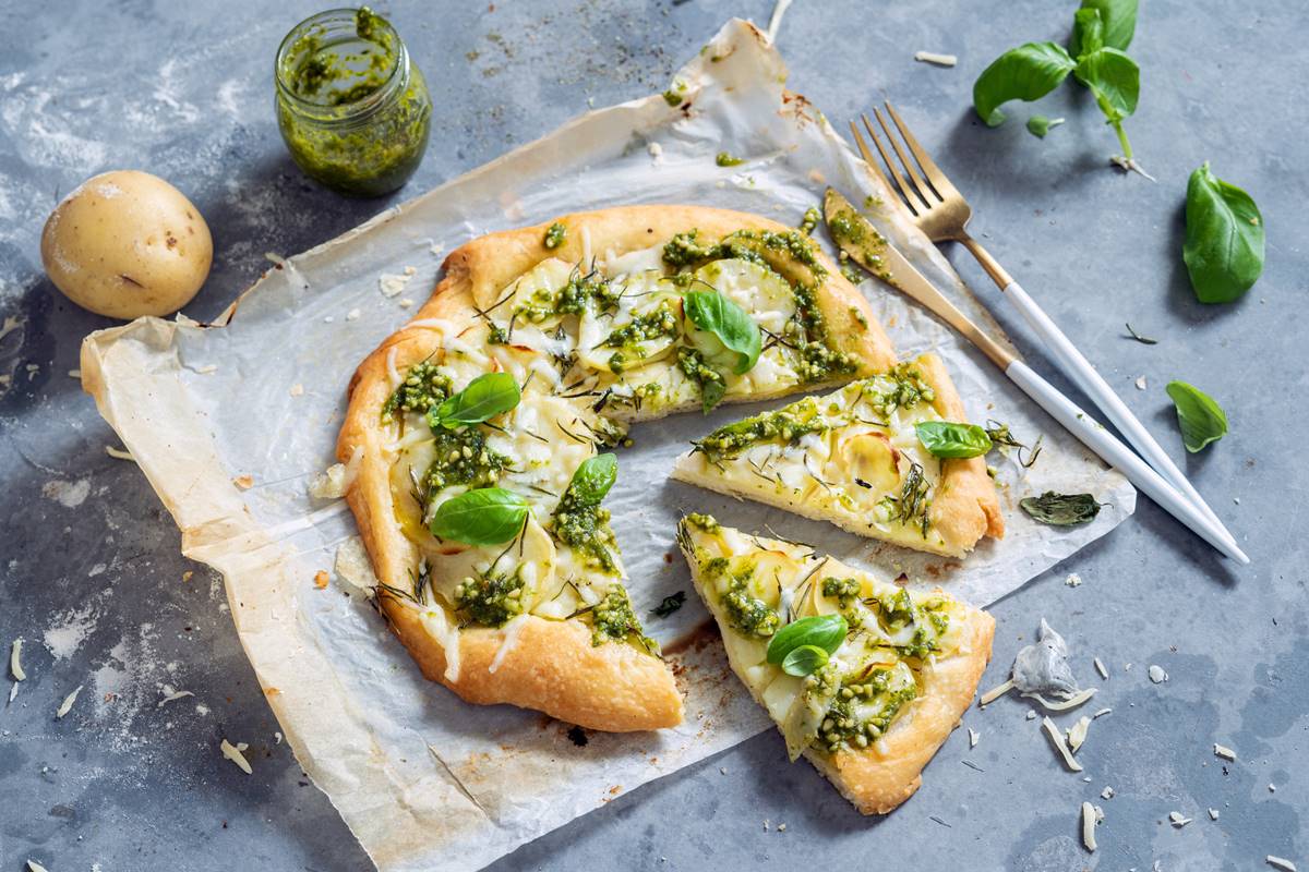 Foto colorida de pizza vegetariana com dois pedaços cortados. Ao lado tem um pote de molho pesto, manjericão e um par de garfo e faca - Metrópoles