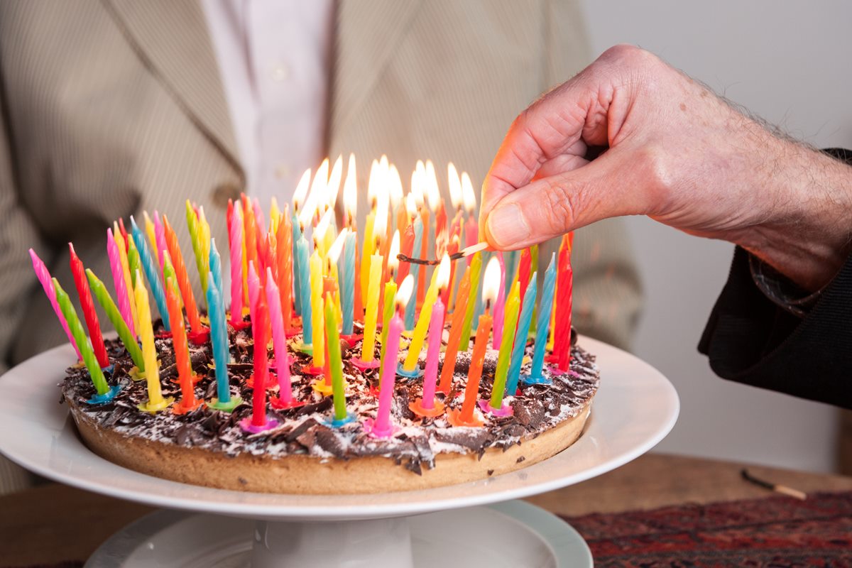 Foto colorida de bolo com velinhas acesas em cima. Uma mão de uma pessoa idosa segura uma das velinhas - Metrópoles