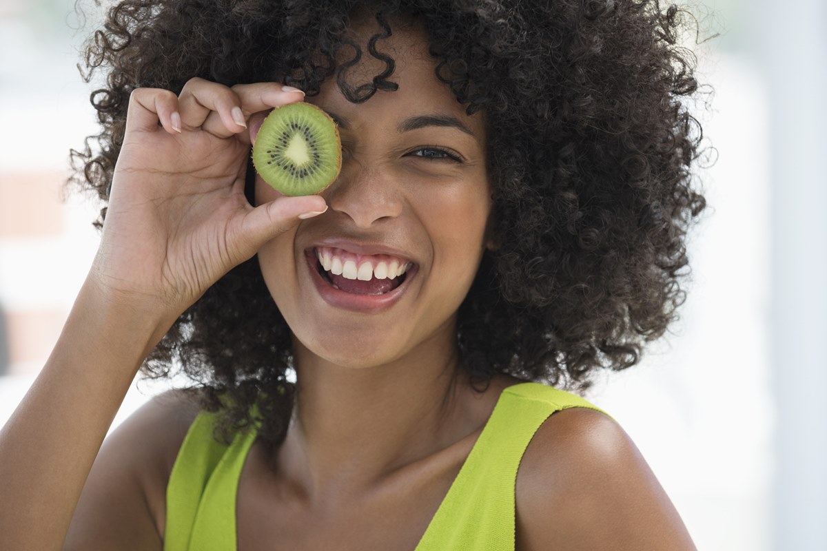 Foto colorida de mulher negra segurando kiwi próximo ao olho. Ela usa uma blusa verde - Metrópoles