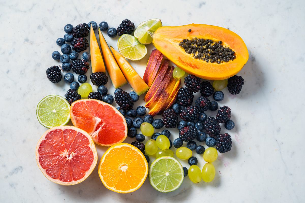 Foto colorida de frutas em cima de uma superfície branca. As frutas são amora, mamão, laranja, limão, uva e cranberry - Metrópoles