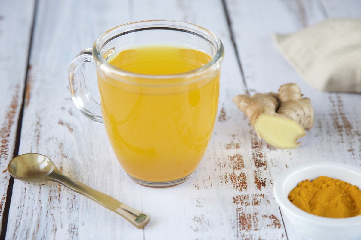 Foto colorida de xícara transparente com chá amarelo. Ao lado do utensílio tem uma colher, raízes de cúrcuma e um pote branco com a especiaria em pó - Metrópoles