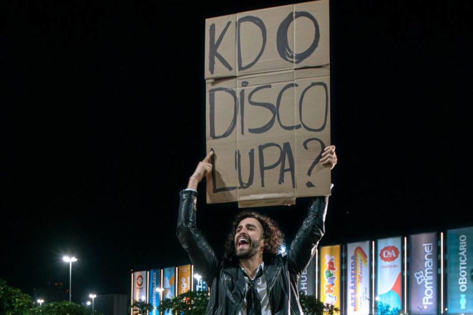 Foto colorida de Múcio Botelho, vocalista da banda Lupa, segurando cartaz - Metrópoles