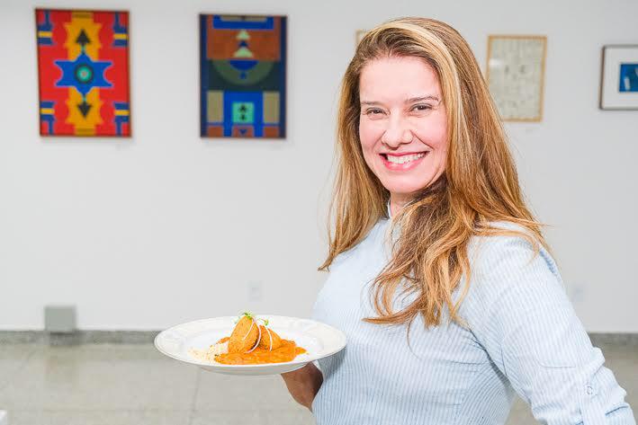 Foto colorida. Chef Leninha Camargo,uma mulher loira e branca, está segurando um prato que preparou olhando sorridente para a câmara - Metrópoles