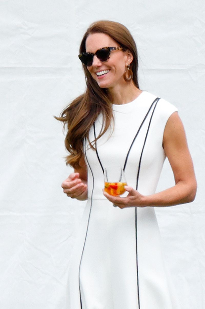 Foto colorida de mulher branca, de cabelo castanho e longo, com vestido branco. Ela usa óculos e está com um copo na mão - Metrópoles