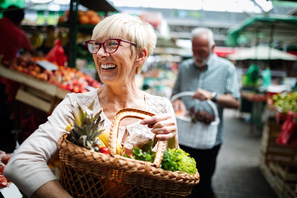 Foto colorida. Mulher idosa branca e de cabelo brancos está segurando uma cesta com vários legumes e verduras - Metrópoles