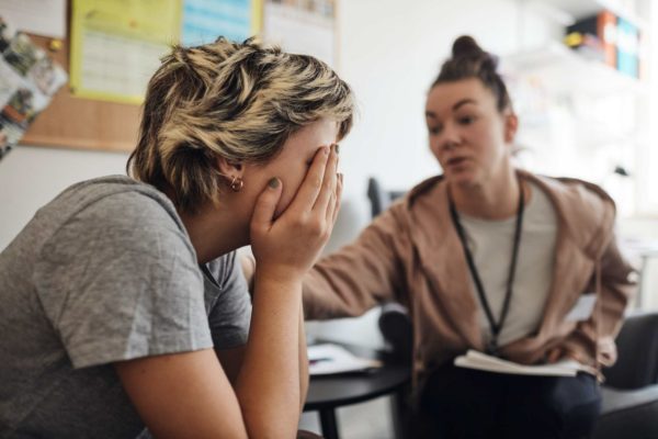 Psicóloga Conselheira não binária consolando aluna cobrindo o rosto com a mão na secretaria da escola