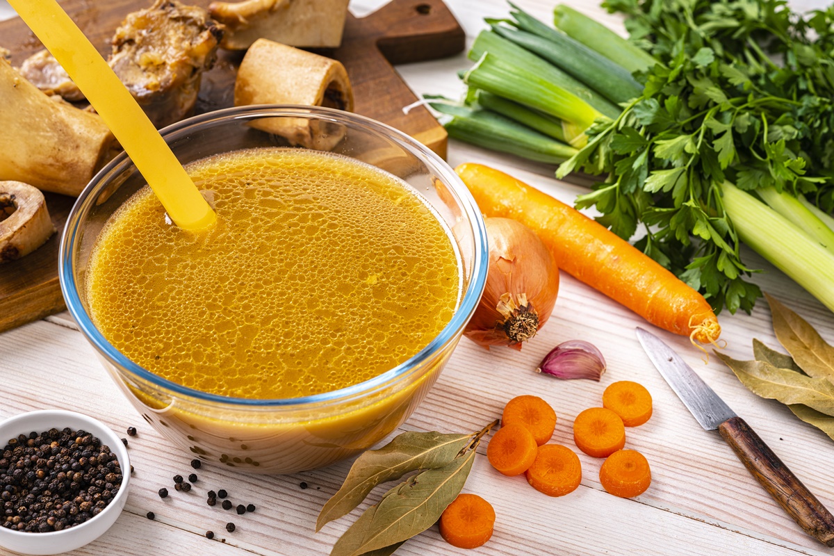 Foto colorida. Vista de alto ângulo de caldo de osso caseiro saudável em uma tigela de vidro na mesa branca. Os ingredientes para cozinhar o caldo de osso estão por todo o recipiente - Metrópoles