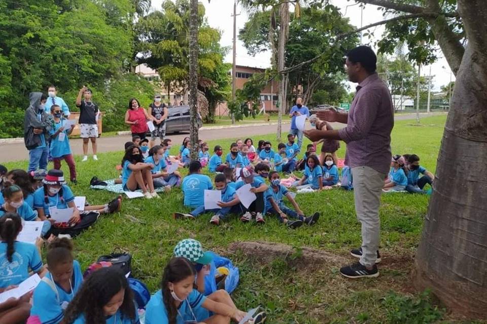 Professor em frente à árvore dando palestra para alunos, sentados na grama