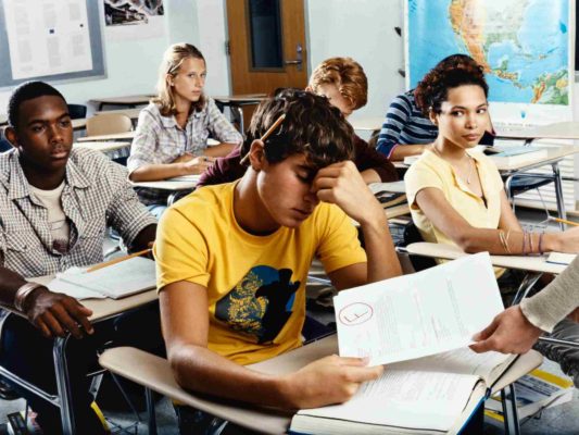 Jovem recebe prova em sala de aula enquanto os colegas o observam