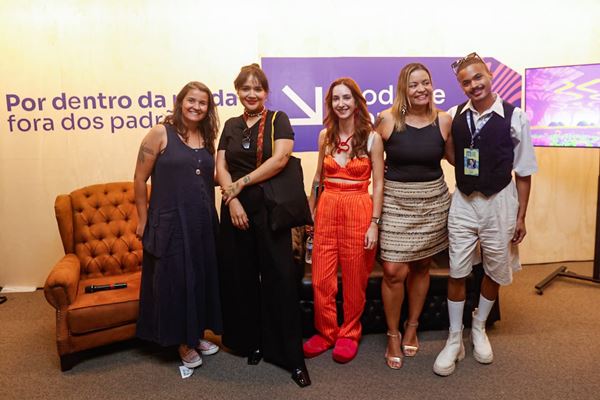 Grupo de pessoas em pé durante evento de moda. Eles estão posicionados em frente a cenário com sofá ao fundo - Metrópoloes