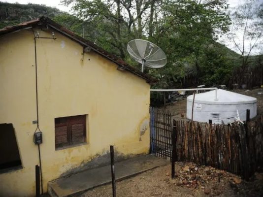 Fotografia de uma casa amarela. Ao lado, uma cisterna construída para captação de água