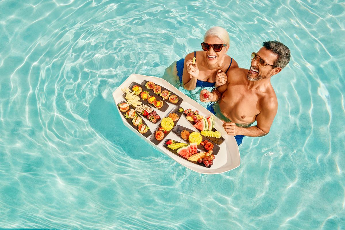 Foto colorida de um casal dentro de uma piscina. À frente deles, há uma canoa com delícias japonesas - Metrópoles 