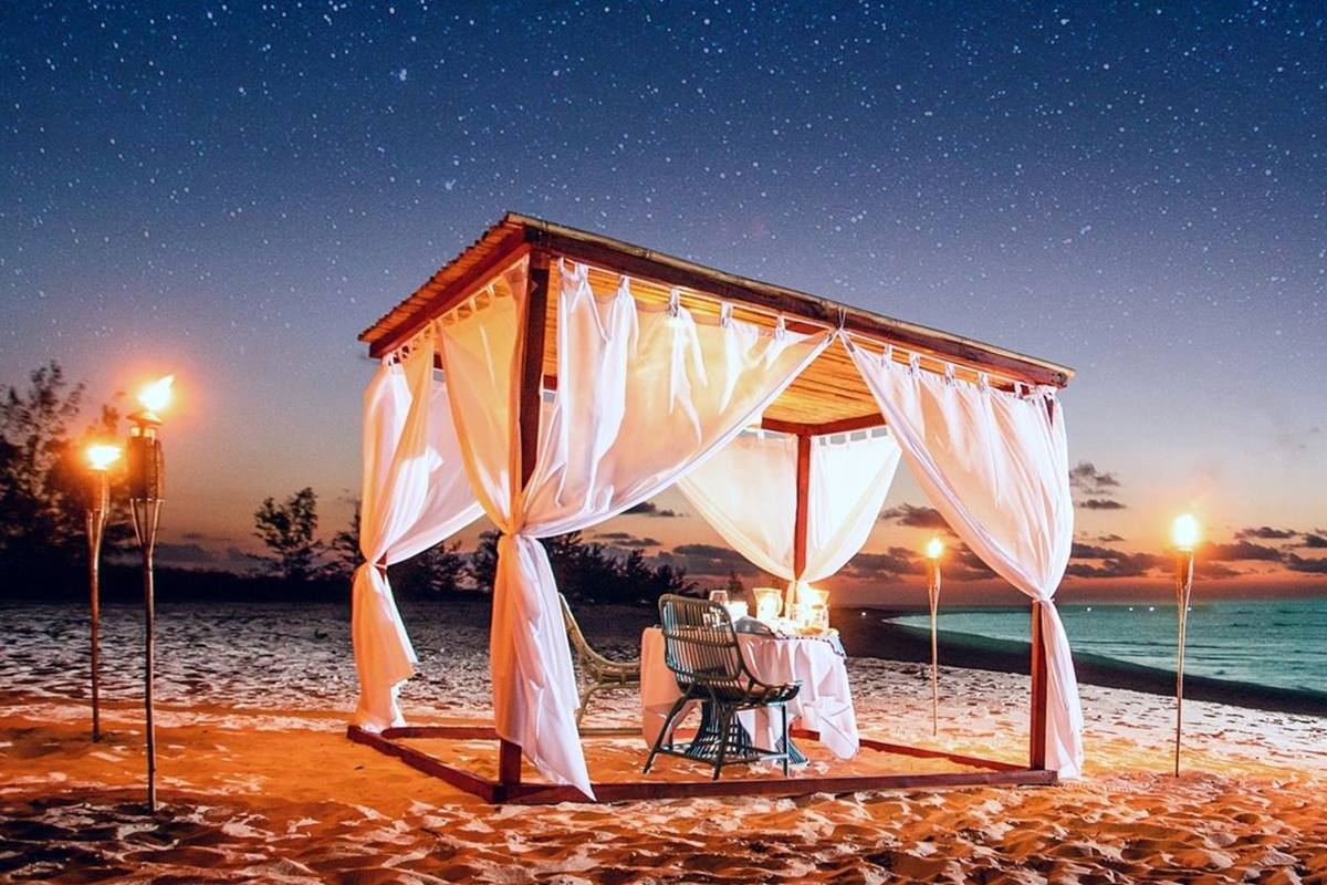 Foto colorida de tenda com mesa para jantar, montada em areia de praia, com tochas ao redor - Metrópoles