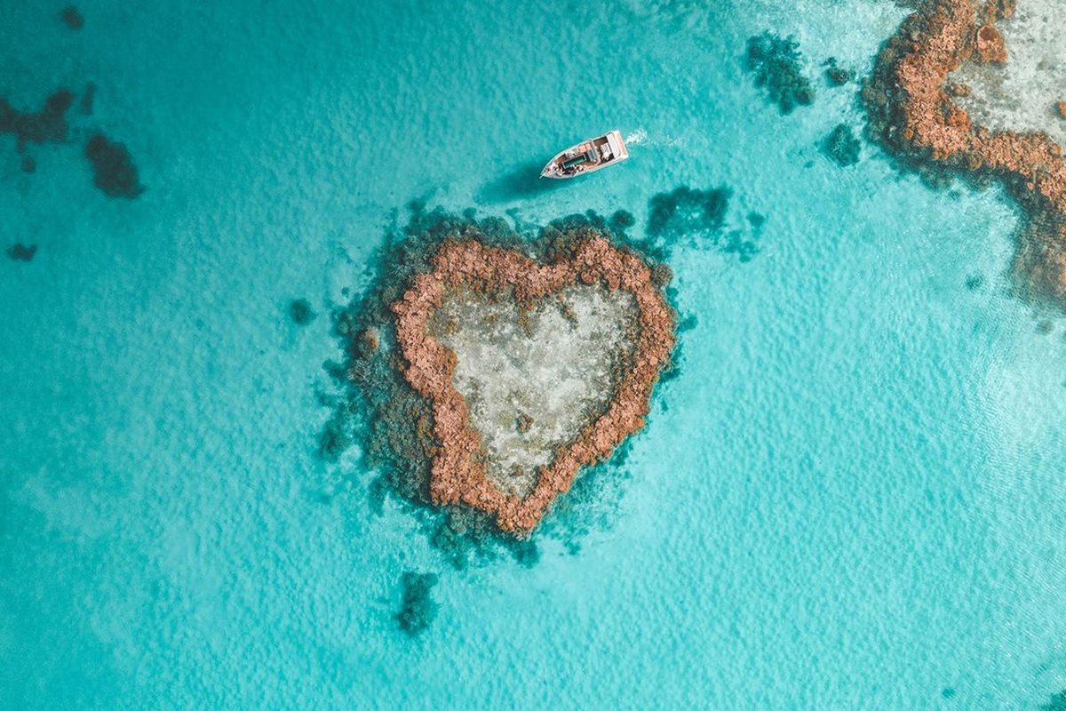 Foto colorida de rocha em formato de coração em mar azul-turquesa com barco passando ao lado - Metrópoles